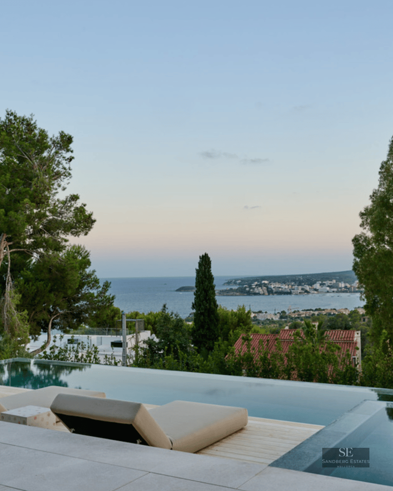 Modern infinity pool with two beige loungers overlooking a calm blue sea and coastal town at dusk.