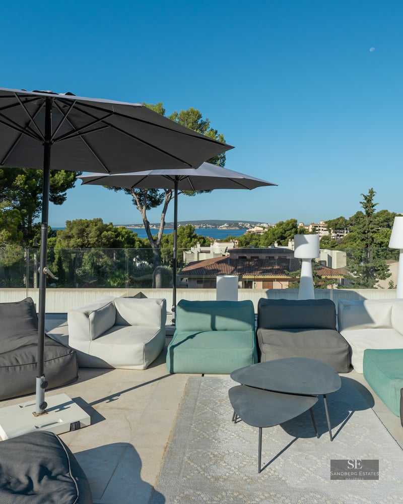 Modern rooftop terrace with grey and green lounge sofas, large black umbrellas, and glass railings overlooking the sea.