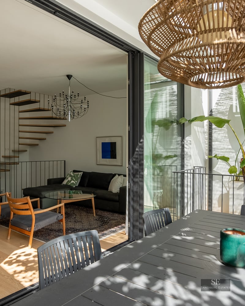 Modern living room with a floating wooden staircase seen through open glass doors from a terrace with tropical plants.