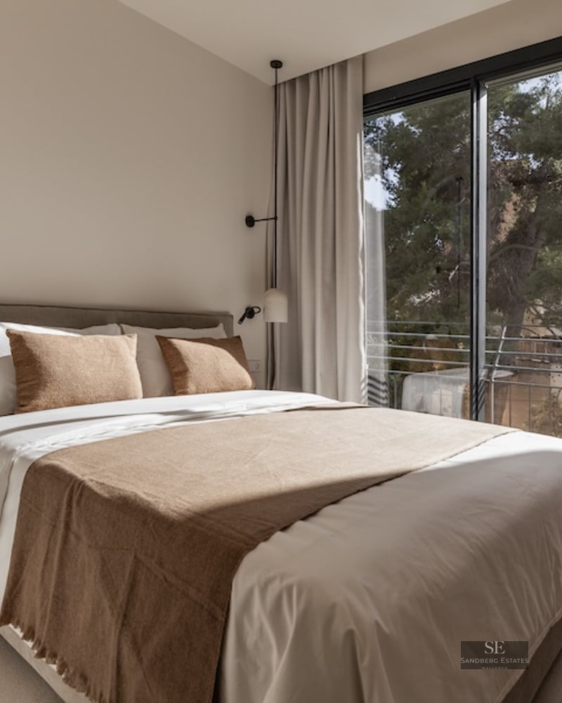 Bright bedroom with a large bed, neutral linens, designer pendant lights, and a floor-to-ceiling window viewing trees.