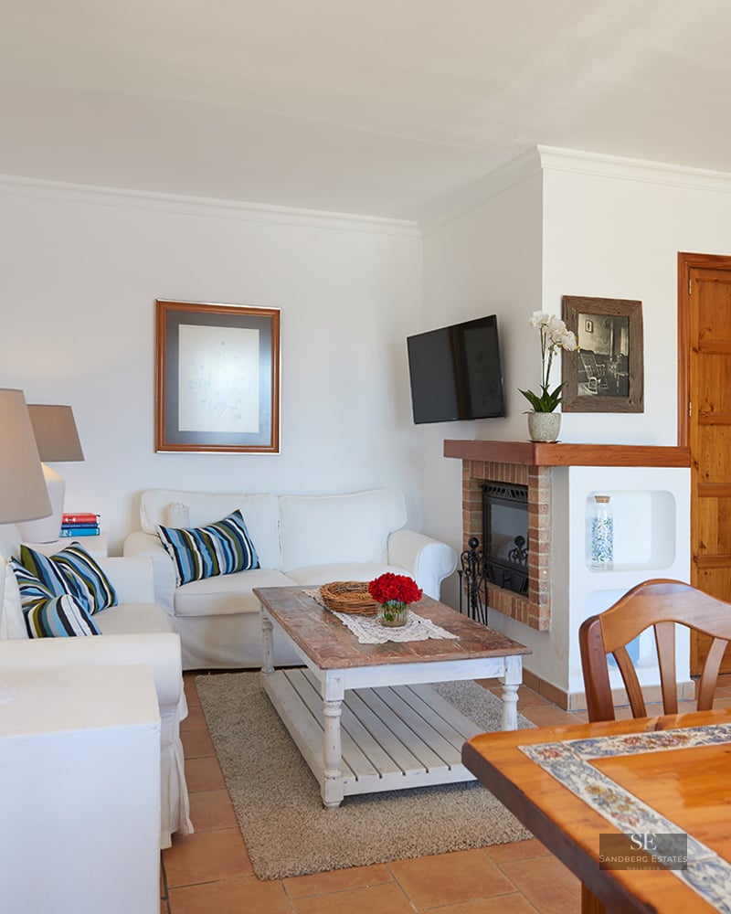 Bright living room with white sofas, a rustic wooden coffee table, a fireplace, and terracotta tile flooring.