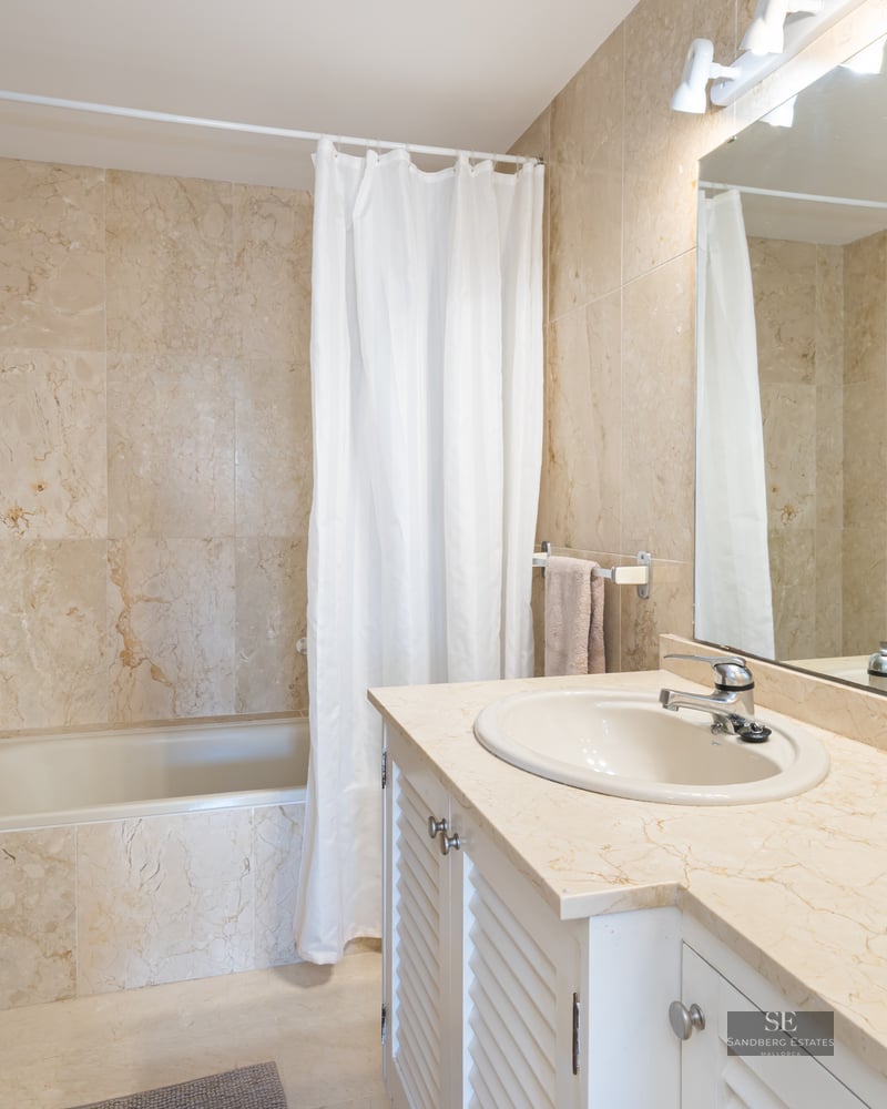 Bathroom featuring beige marble walls, a large mirror, white vanity with marble top, and a bathtub.