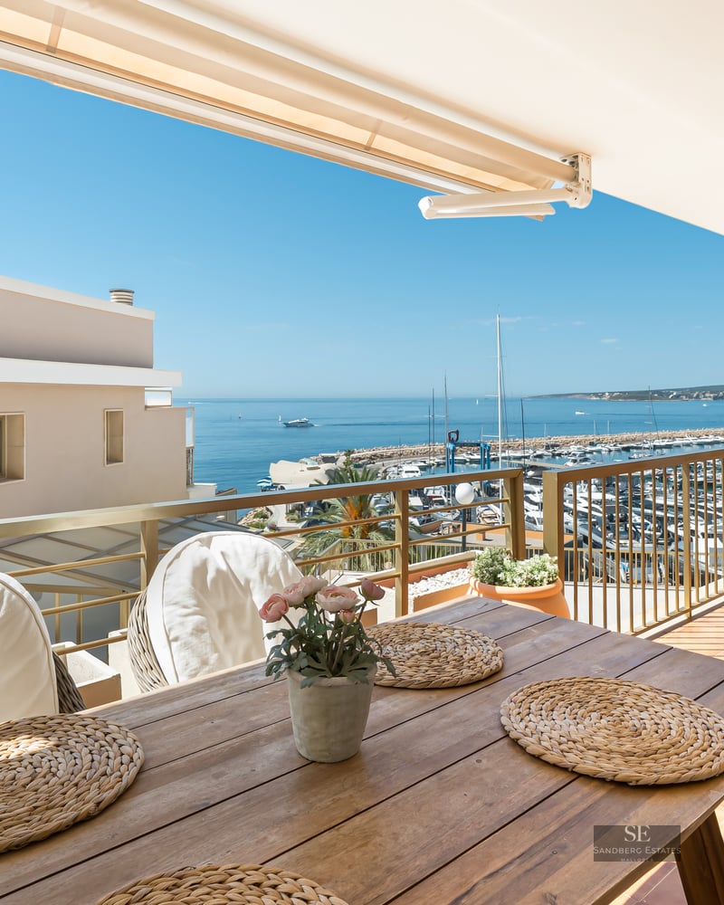 Balcony with wooden dining table and white chairs overlooking a marina and blue sea under a clear sky.