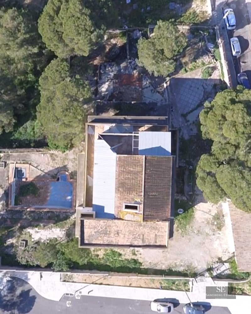 Top-down aerial view of a house with tiled roof, empty swimming pool, and surrounding pine trees.