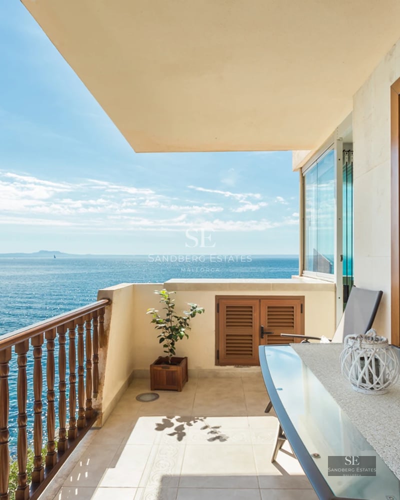 A sunny balcony with a glass dining table and wooden railing overlooking a vast blue ocean under a clear sky.