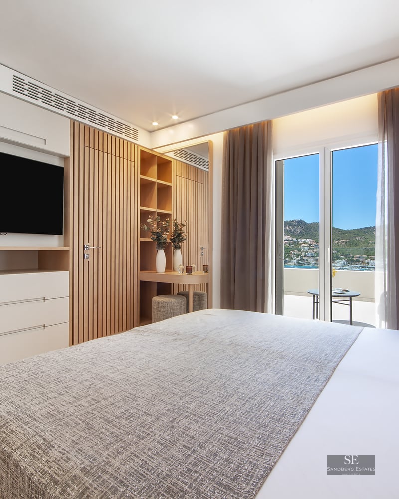 Bright bedroom with large bed, wood slat panels, wall-mounted TV, and balcony overlooking coast and mountains.