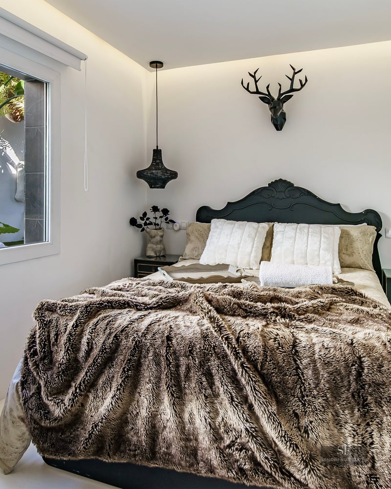 Bedroom with a brown faux fur throw, dark headboard, deer head wall sculpture, and window looking onto tropical plants.