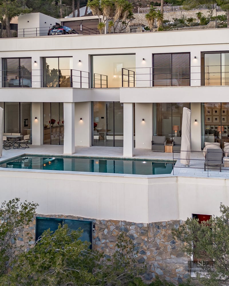 Two-story modern white villa with an infinity pool on a stone-clad terrace overlooking a hillside.