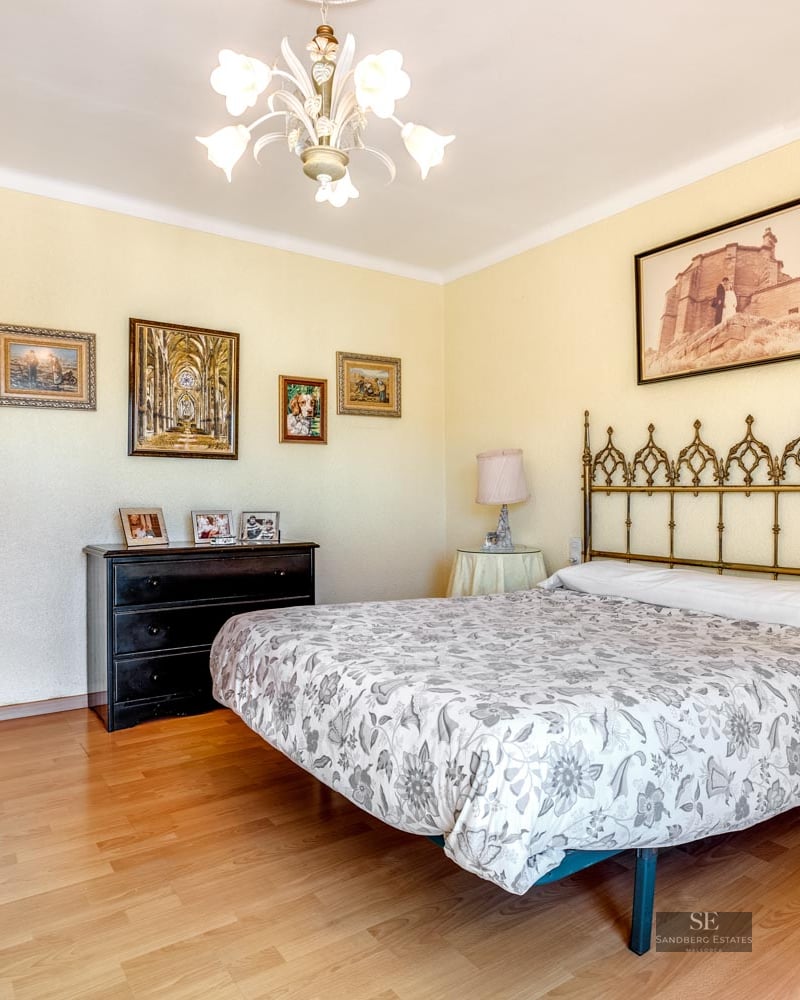 A bedroom with a floral bed, metal headboard, wooden floors, yellow walls, and framed pictures.
