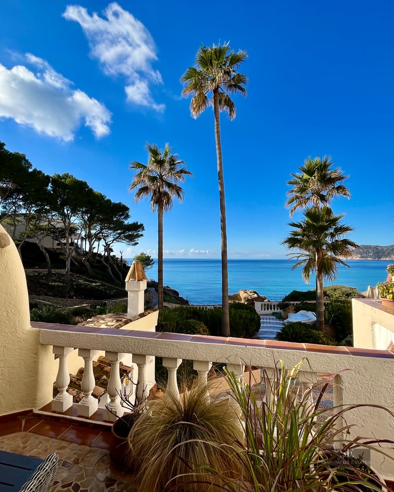 Terrace with a white built-in outdoor fireplace and balustrade overlooking palm trees and the vibrant blue sea.