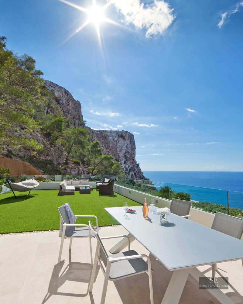 Modern outdoor terrace with dining area, artificial lawn, and glass railing overlooking the sea and cliffs.