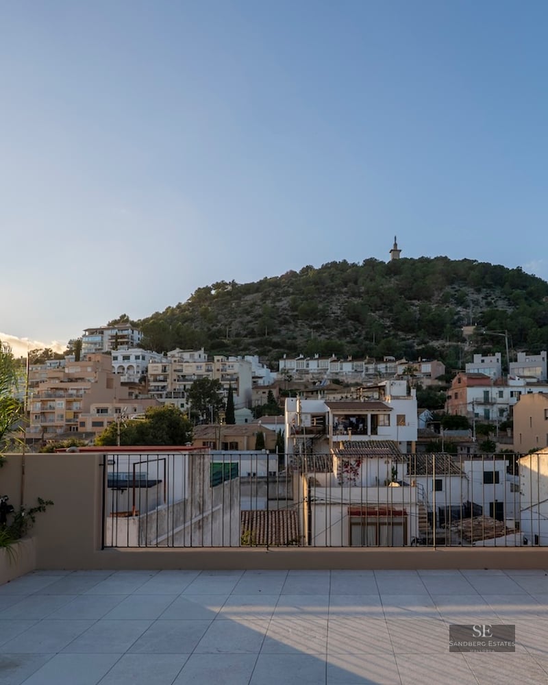 Grande terrasse carrelée avec garde-corps métallique offrant une vue sur un village méditerranéen et une colline au coucher du soleil.