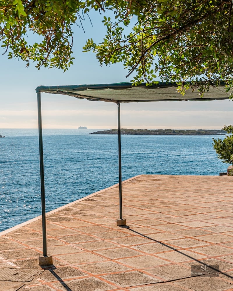A sun-drenched stone terrace with a shade structure overlooking the sparkling blue ocean and a distant island.