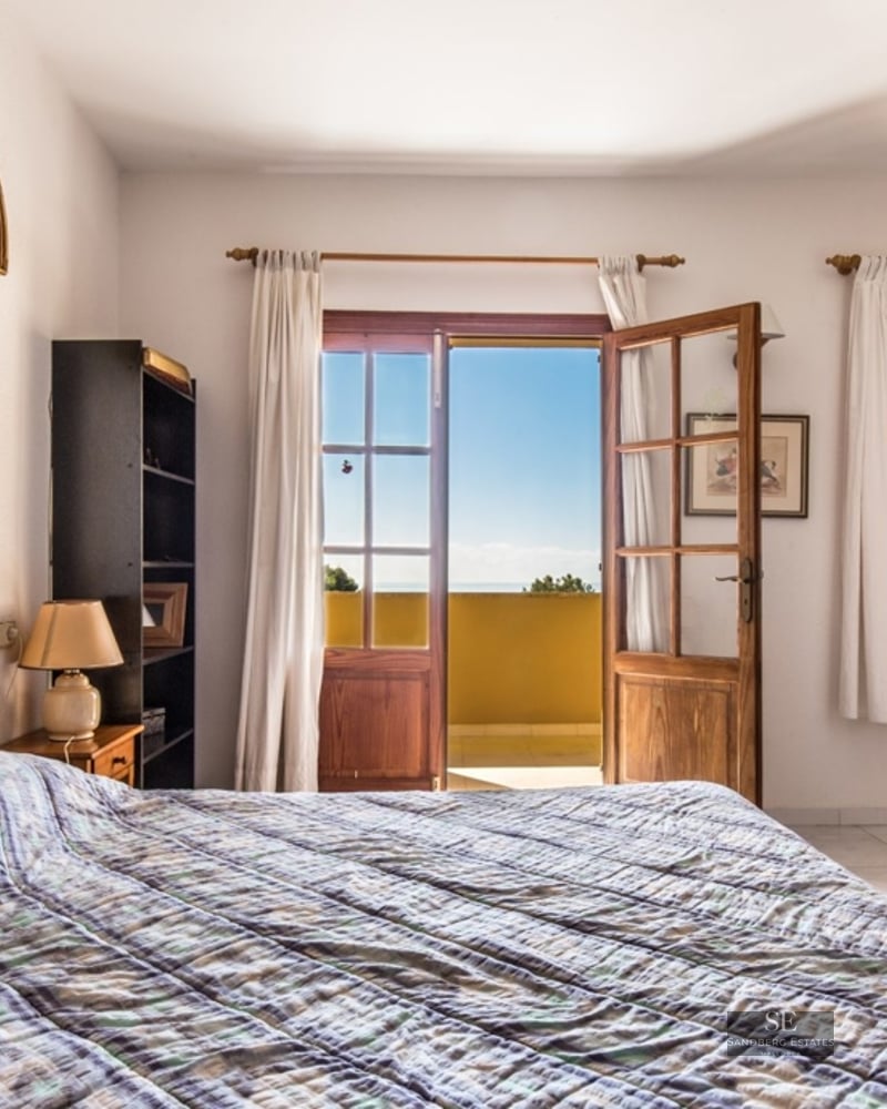 A bright bedroom with a double bed, white textured walls, and open wooden doors leading to a sunny balcony with a view.
