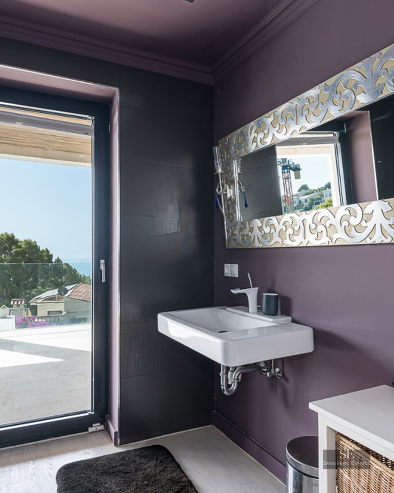 Modern bathroom with plum-colored walls, floating white sink, ornate silver mirror, and glass door to a sunny terrace.