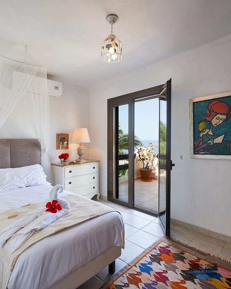A white bedroom with a mosquito net over the bed, a colorful rug, and open glass doors leading to a sunny terrace.