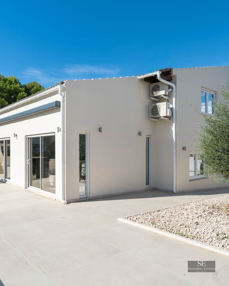 Grande terrasse en pierre blanche avec table à manger, olivier et façade de villa moderne sous un ciel bleu dégagé.
