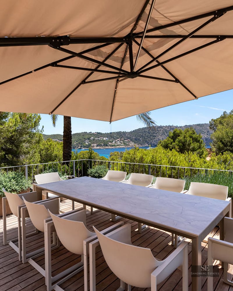 Outdoor wooden terrace with a dining table and chairs under a large umbrella overlooking a sea bay.