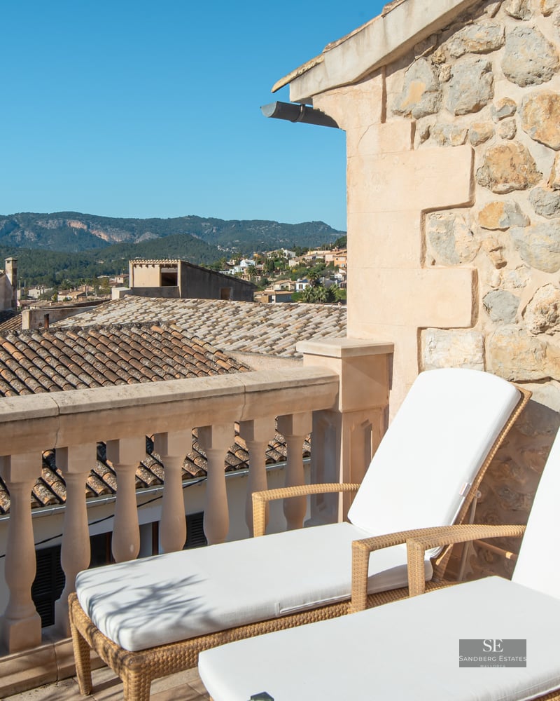 Two white sun loungers on a stone terrace overlooking a Mediterranean village and mountains.