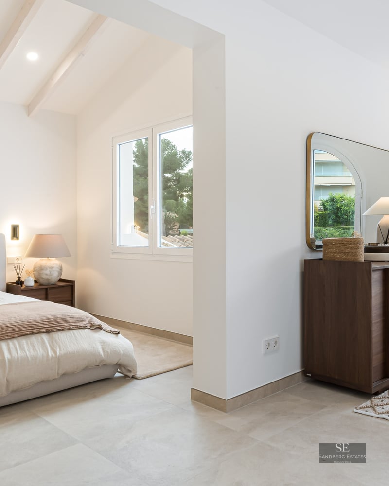 Modern master bedroom with white beamed ceilings, king bed, dark wood dresser, and natural light from large windows.