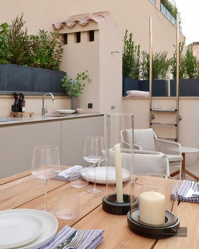 Wooden dining table on a modern terrace featuring an outdoor kitchen and lush greenery.