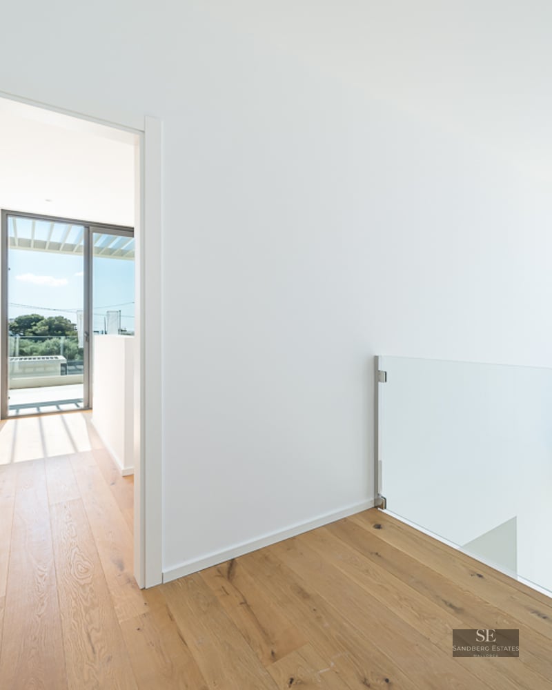 Bright hallway with light wood flooring, glass railing, and views through large windows to a sunny exterior.