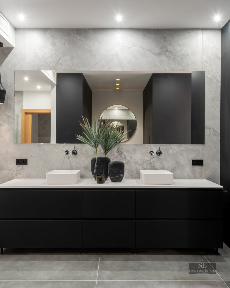 Luxury bathroom with marble walls, double white basins on a black vanity unit, and a large mirror.
