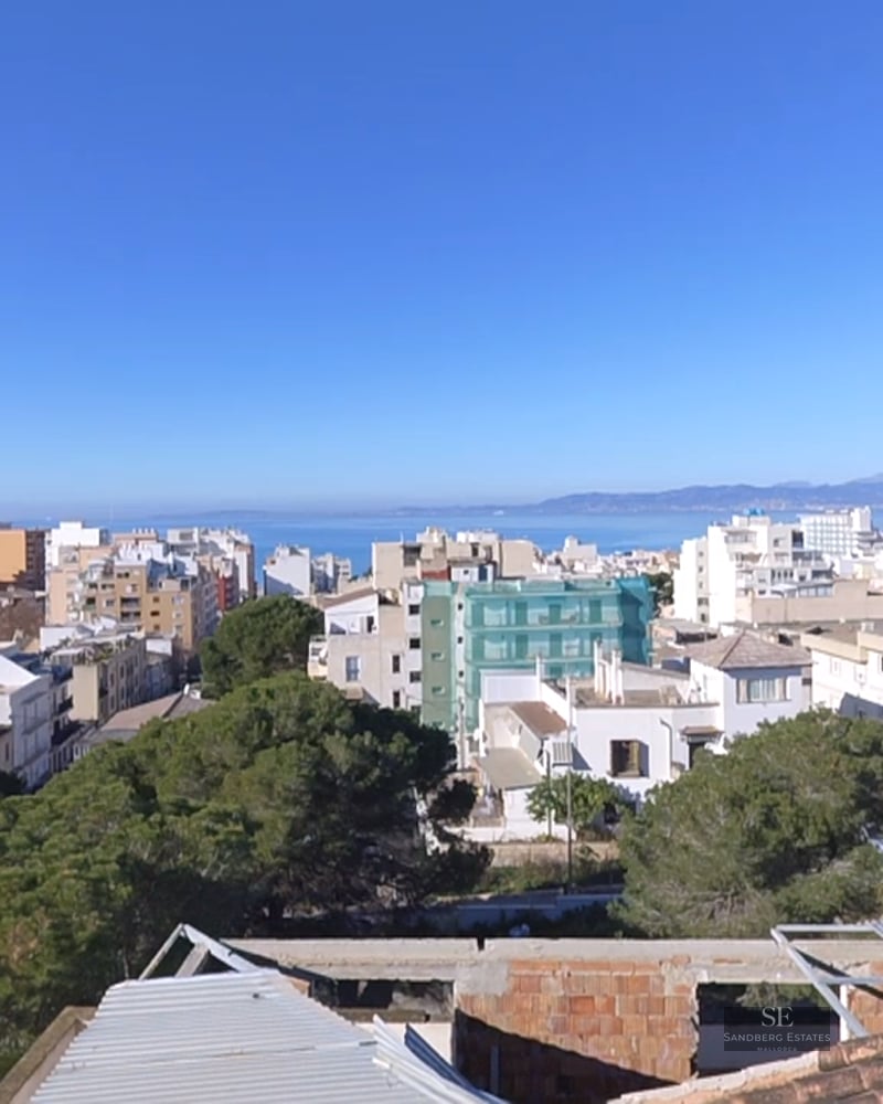 High-angle view of a coastal city with white buildings, green pine trees, and the blue Mediterranean sea in the background.