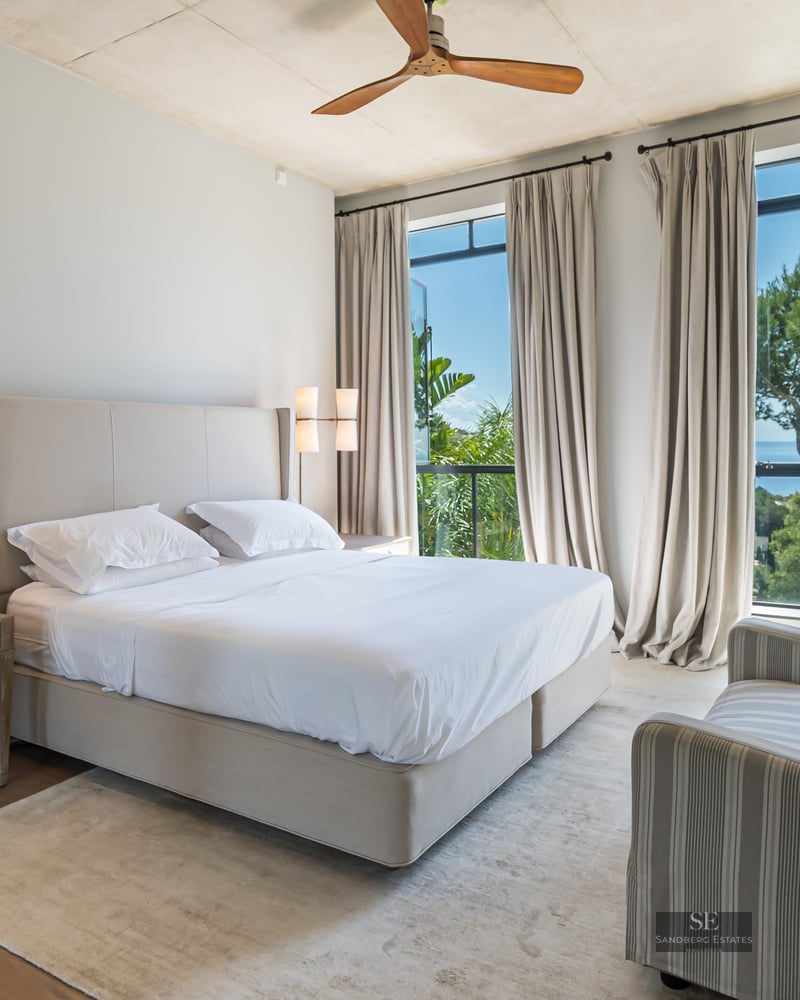 Bright master bedroom with a large bed, striped sofa, and floor-to-ceiling windows overlooking lush trees and the sea.