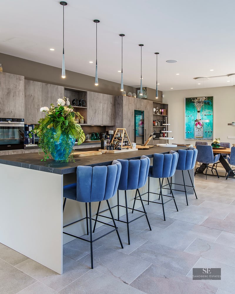 Cuisine moderne avec îlot central, tabourets en velours bleu et salle à manger intégrée baignée de lumière.