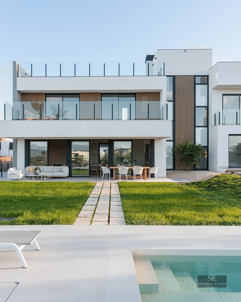 Modern white two-story villa with a turquoise swimming pool, sun lounger, and a green lawn under a clear sky.