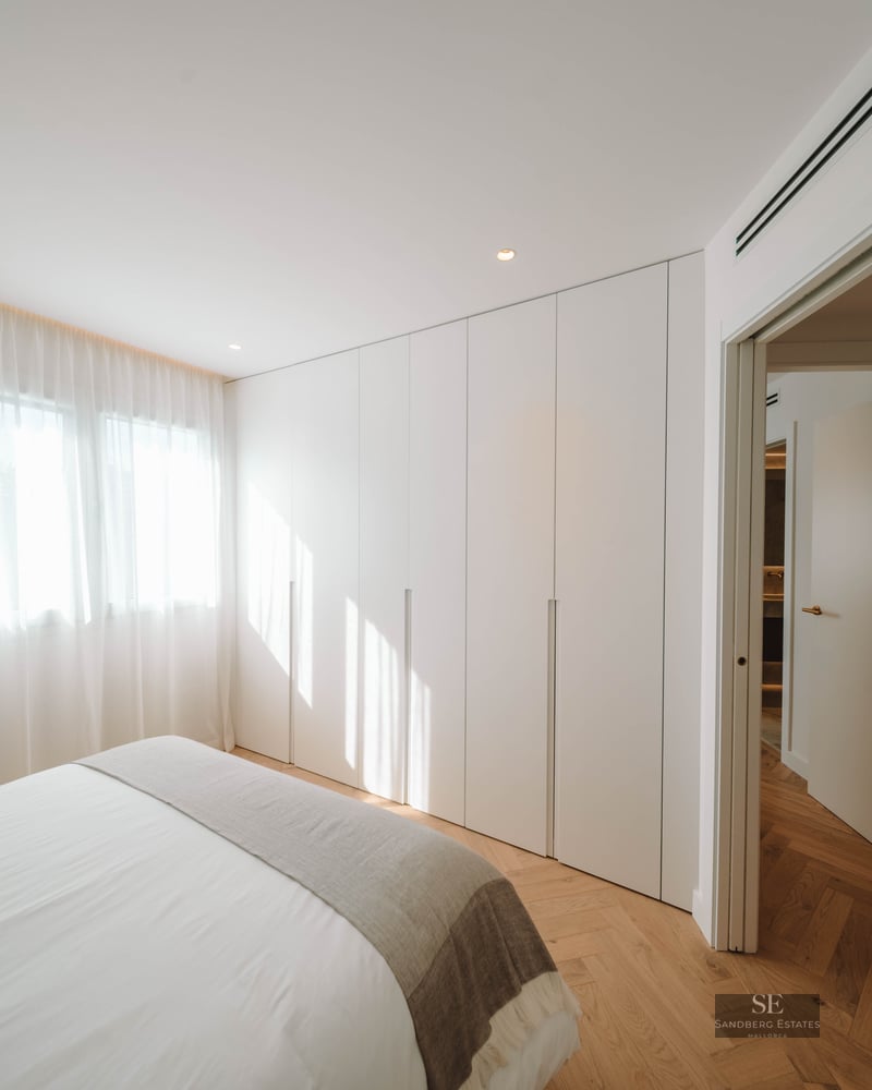 Bright bedroom featuring herringbone wood floors, floor-to-ceiling white wardrobes, a bed with white linens, and sheer curtains.