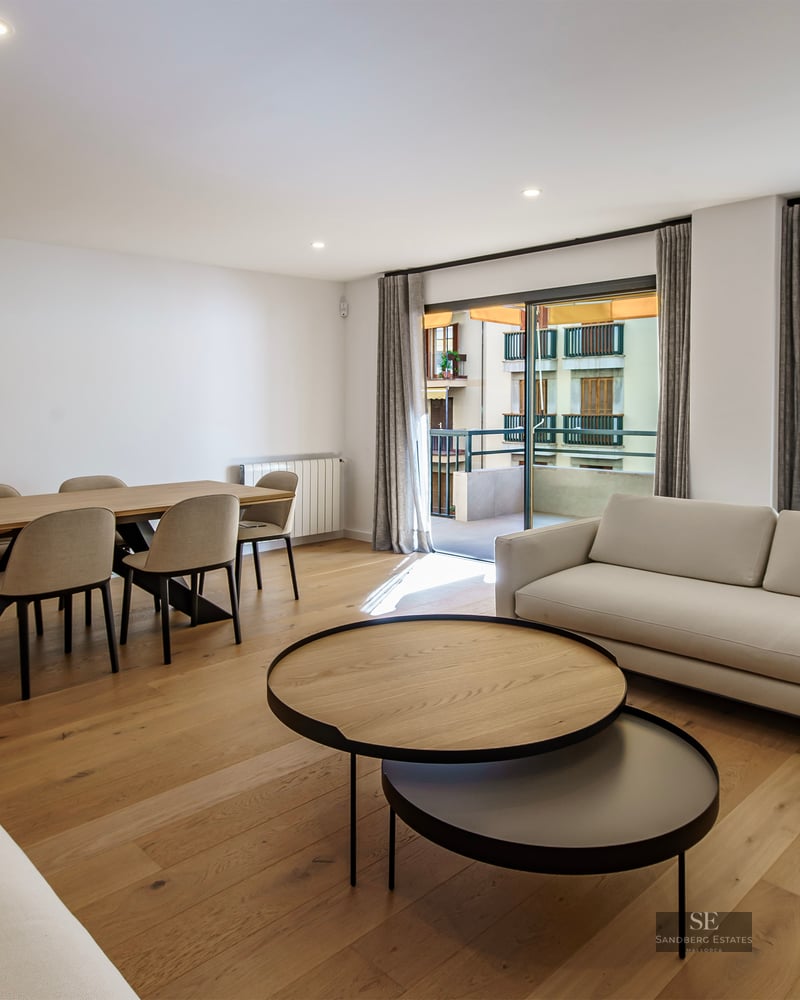 Salon moderne avec canapés blancs, tables basses en bois, coin repas et grandes baies vitrées ouvrant sur un balcon.