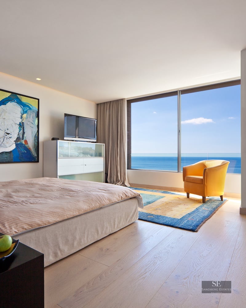 Modern master bedroom featuring floor-to-ceiling sea views, a yellow armchair, and eclectic art.