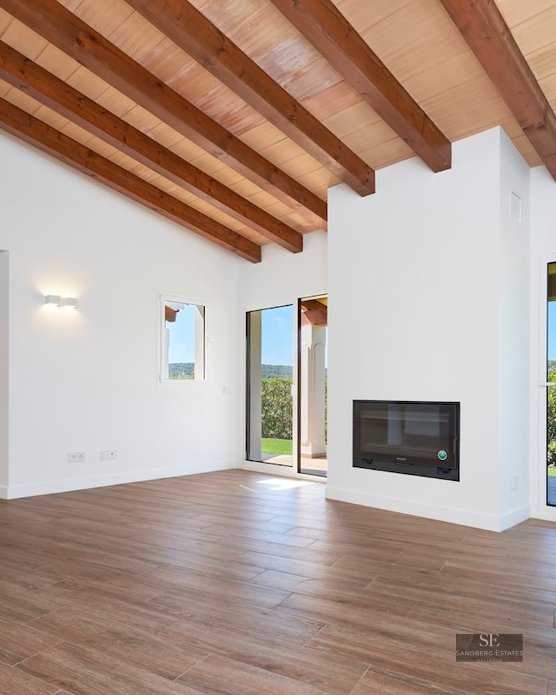 Bright, empty living room featuring exposed wooden ceiling beams, a built-in fireplace, and large glass doors to a garden.