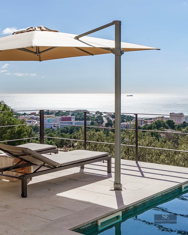 Modern pool terrace with two sun loungers under an umbrella, overlooking the sea and a coastal town.