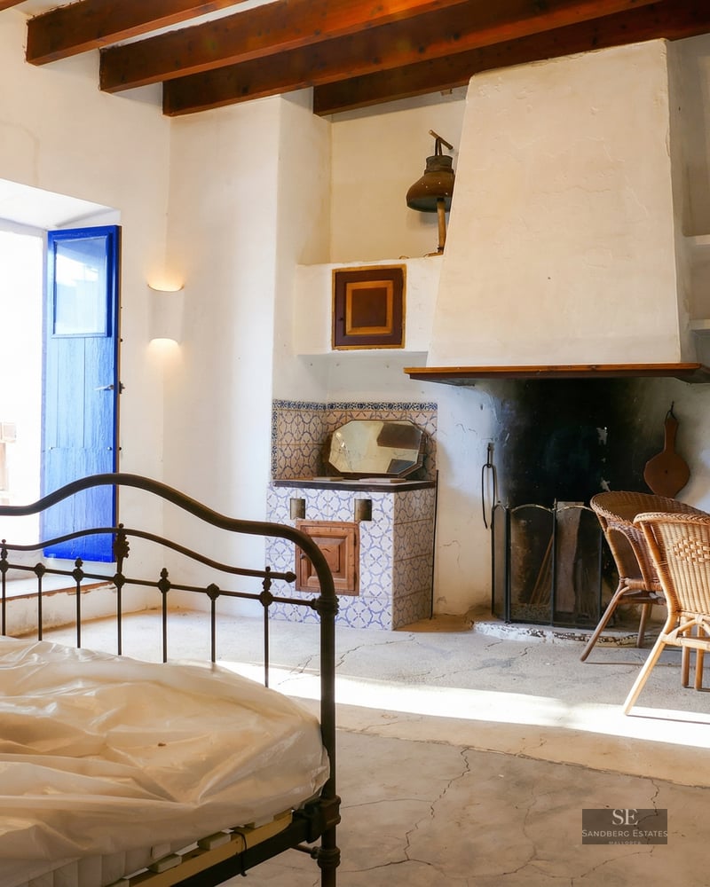 Bedroom with a wrought iron bed frame, a large white fireplace, blue wooden shutters, and wicker chairs under wooden beams.