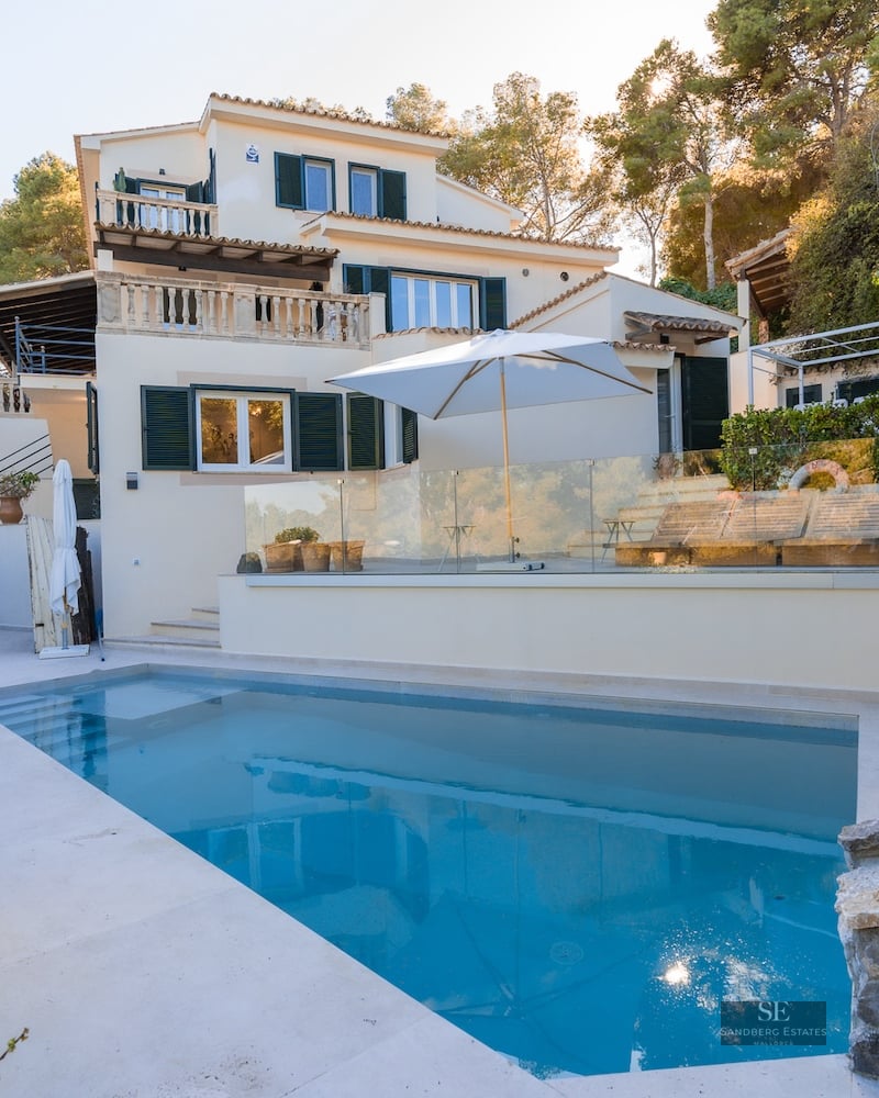 Rectangular swimming pool in the foreground of a multi-level white Mediterranean villa with green shutters and lush trees.