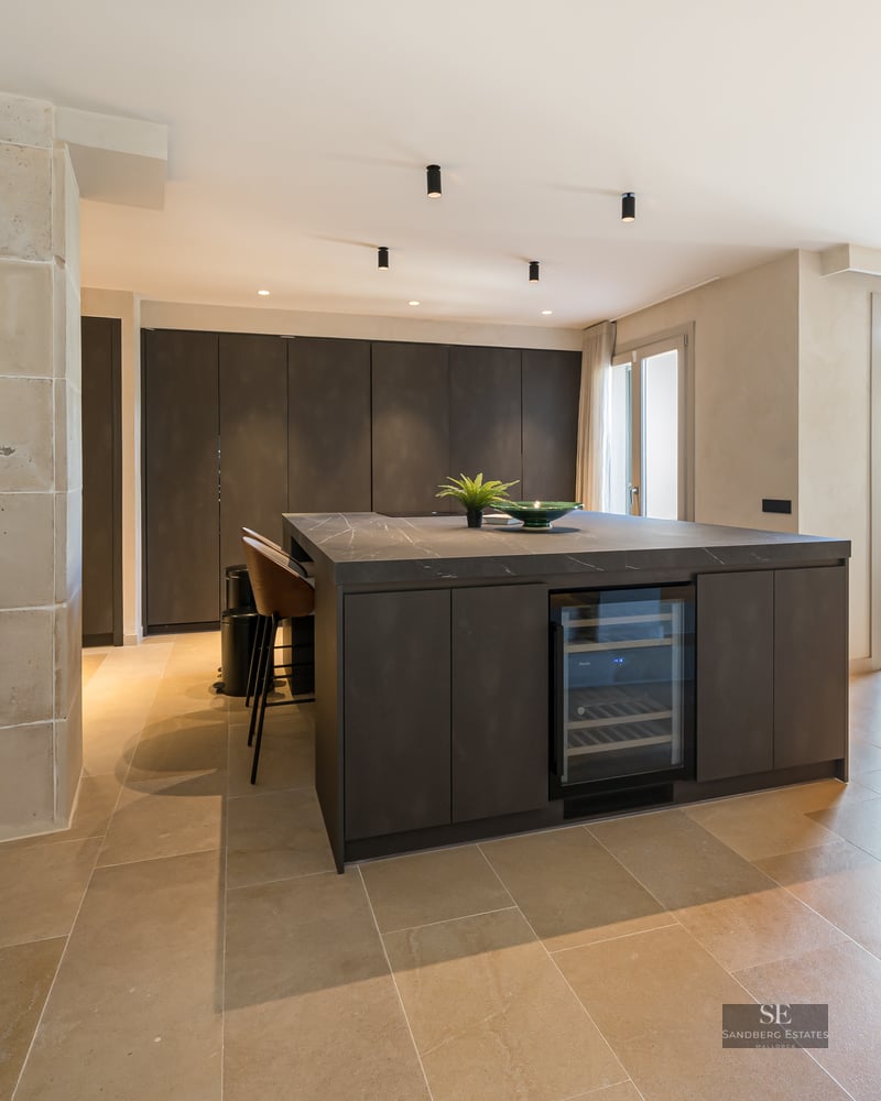 Modern kitchen with dark island and wine fridge next to a rustic stone arch and open-plan dining area.