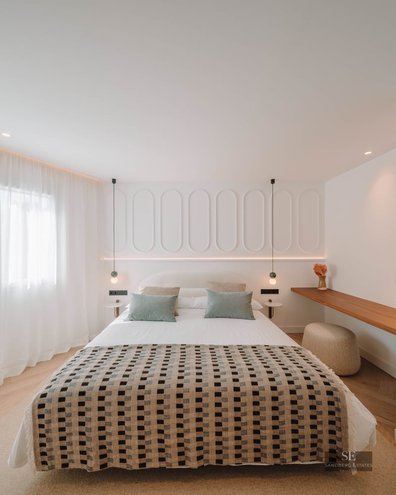 A bright, modern bedroom featuring a patterned bedspread, white paneled headboard wall, and herringbone wood flooring.