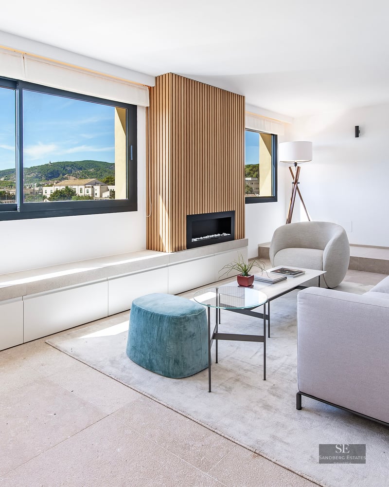 Bright living room featuring a grey sofa, teal ottoman, wood slat fireplace wall, and large window overlooking hills.