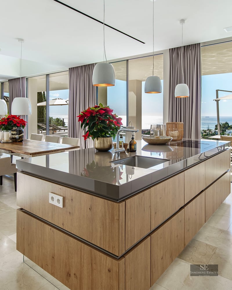Modern kitchen featuring a wooden island, marble floors, and floor-to-ceiling glass walls overlooking the sea.