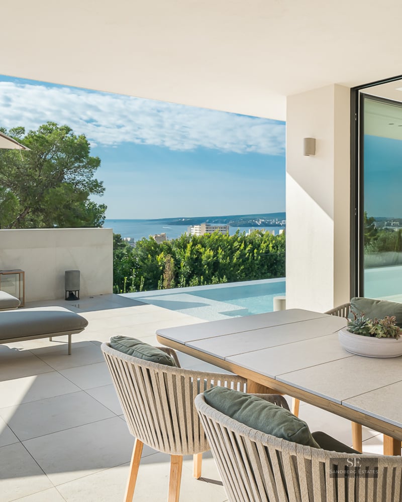 Modern terrace with outdoor dining, lounge chairs, and an infinity pool overlooking the Mediterranean sea and trees.