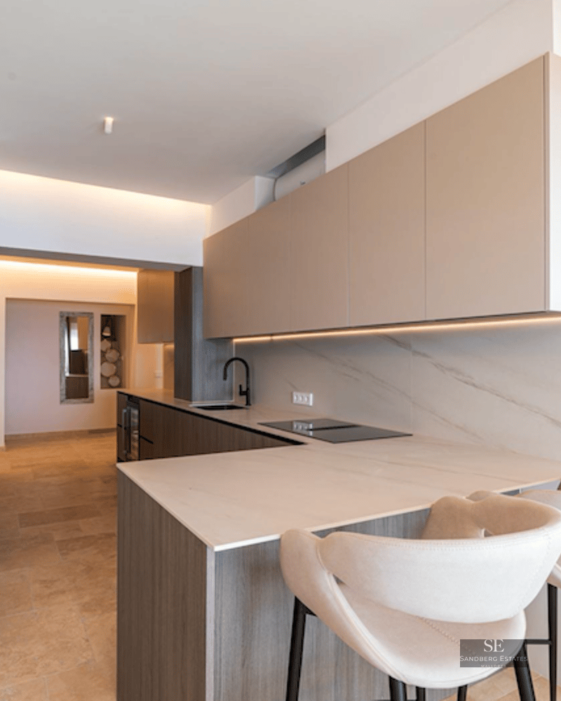 A sleek modern kitchen with light wood cabinets, stone countertops, and cream-colored designer bar stools.