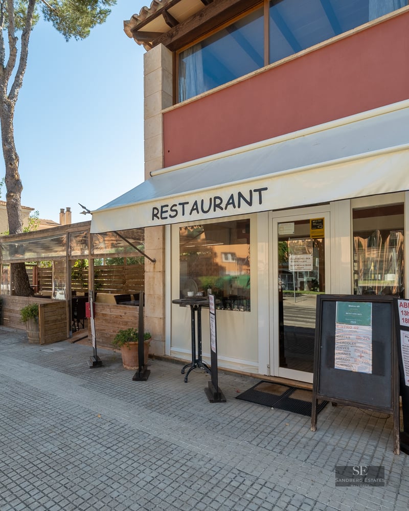 Exterior view of a restaurant with stone walls, white awnings, and a tree-lined sidewalk under a clear sky.