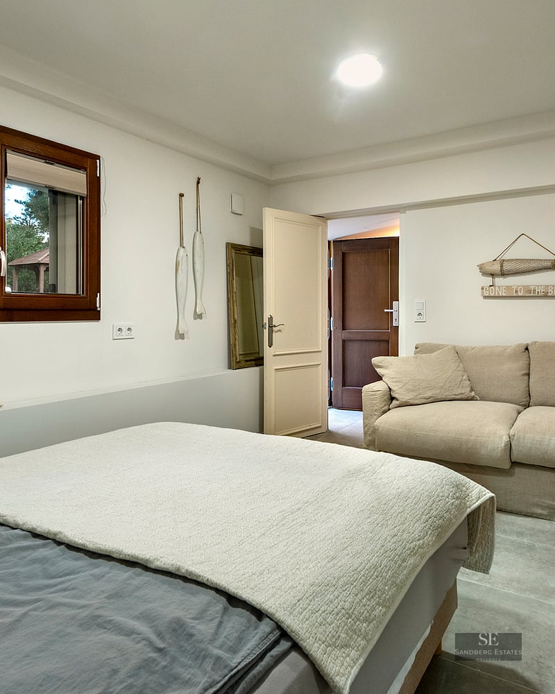 A bright bedroom featuring a grey bed, a beige sofa, a wooden window, and nautical-themed wall decor.