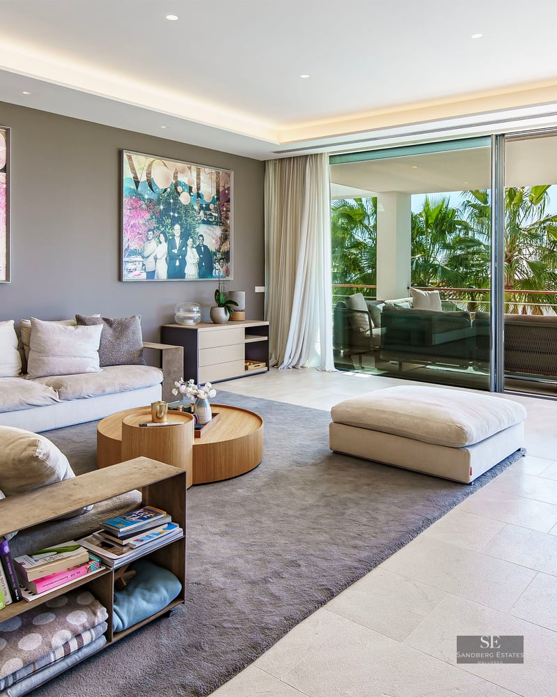 Salon contemporain spacieux avec canapés, art moderne et baies vitrées s'ouvrant sur une terrasse avec palmiers.