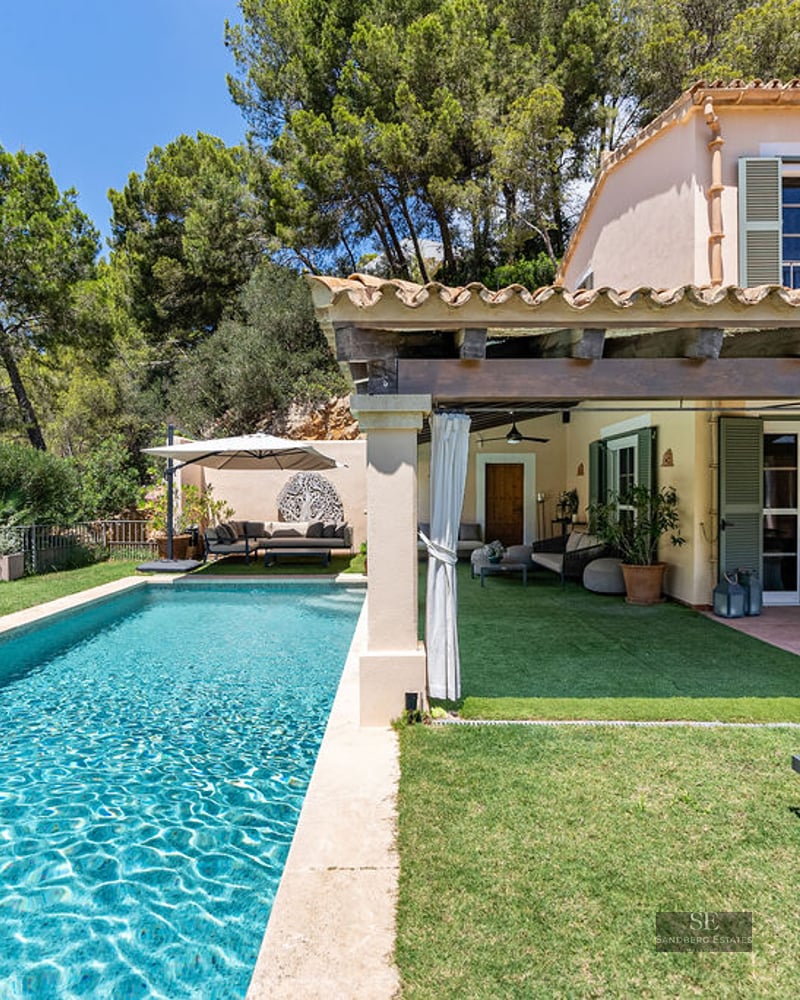 Rectangular turquoise pool next to a Mediterranean villa with a covered terrace and green shutters under a blue sky.