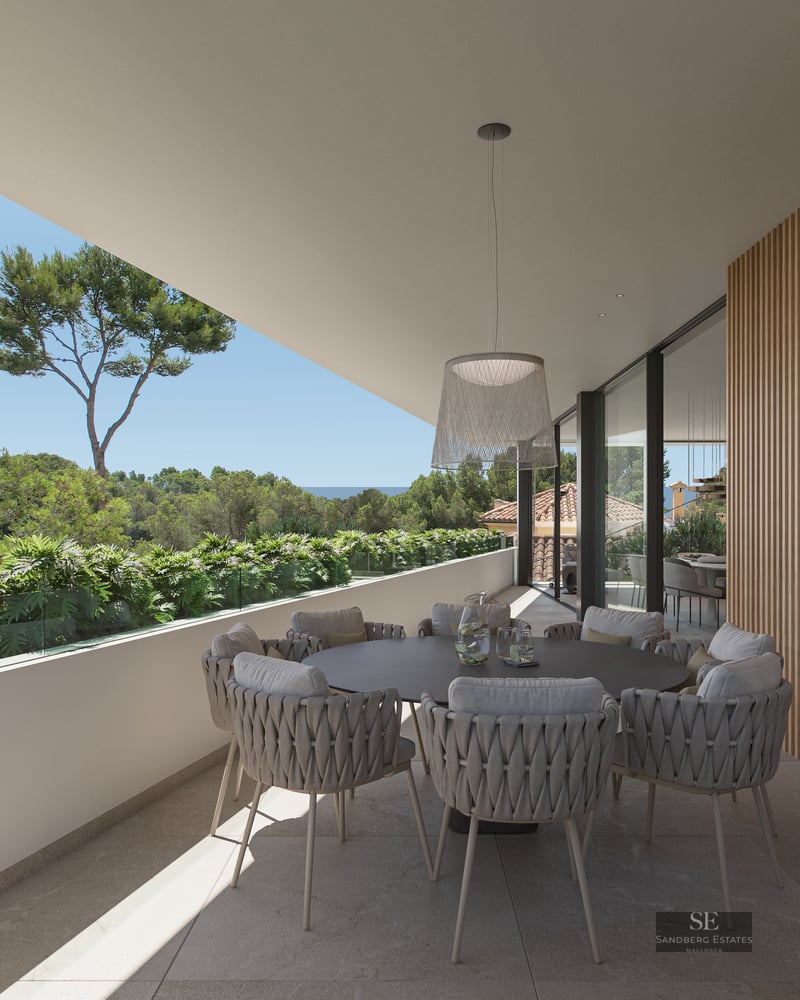 Modern terrace with a round dining table, woven chairs, wood-slatted wall, and a view of lush greenery and the sea.