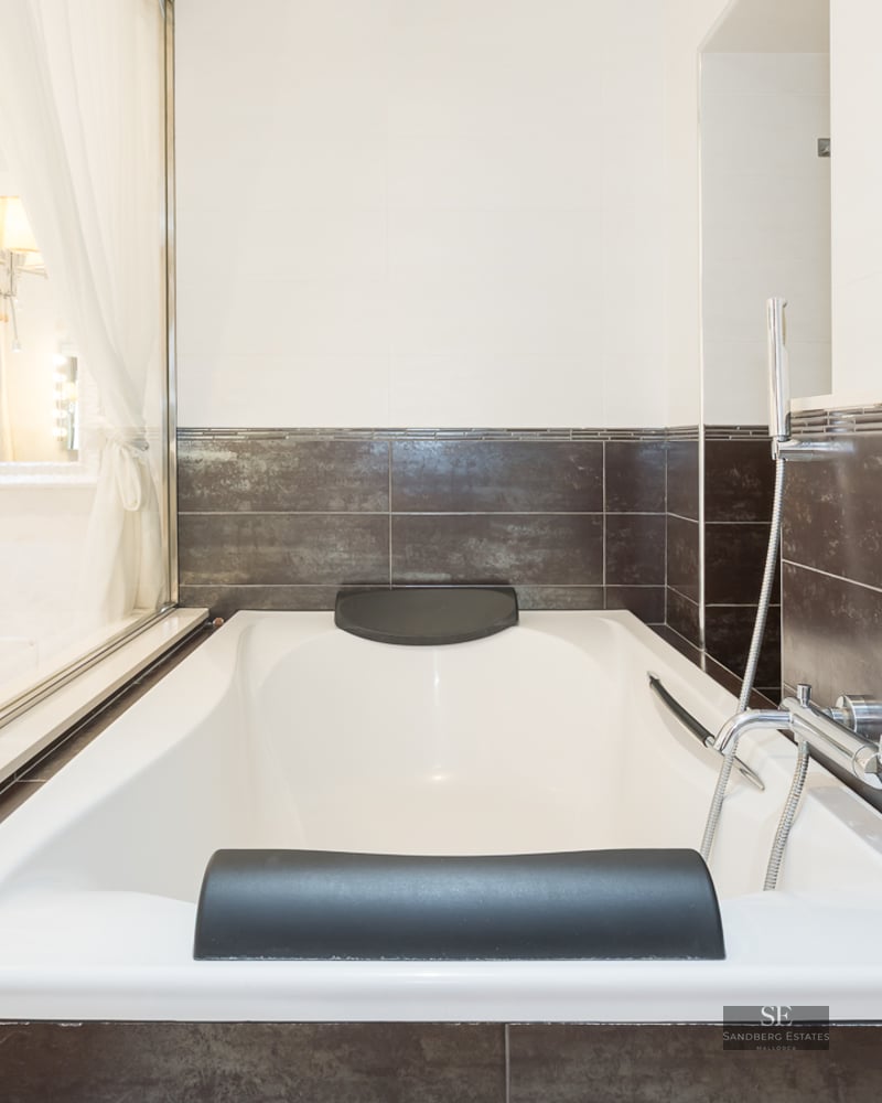 White built-in bathtub with black headrests and dark brown wall tiles in a modern bathroom.